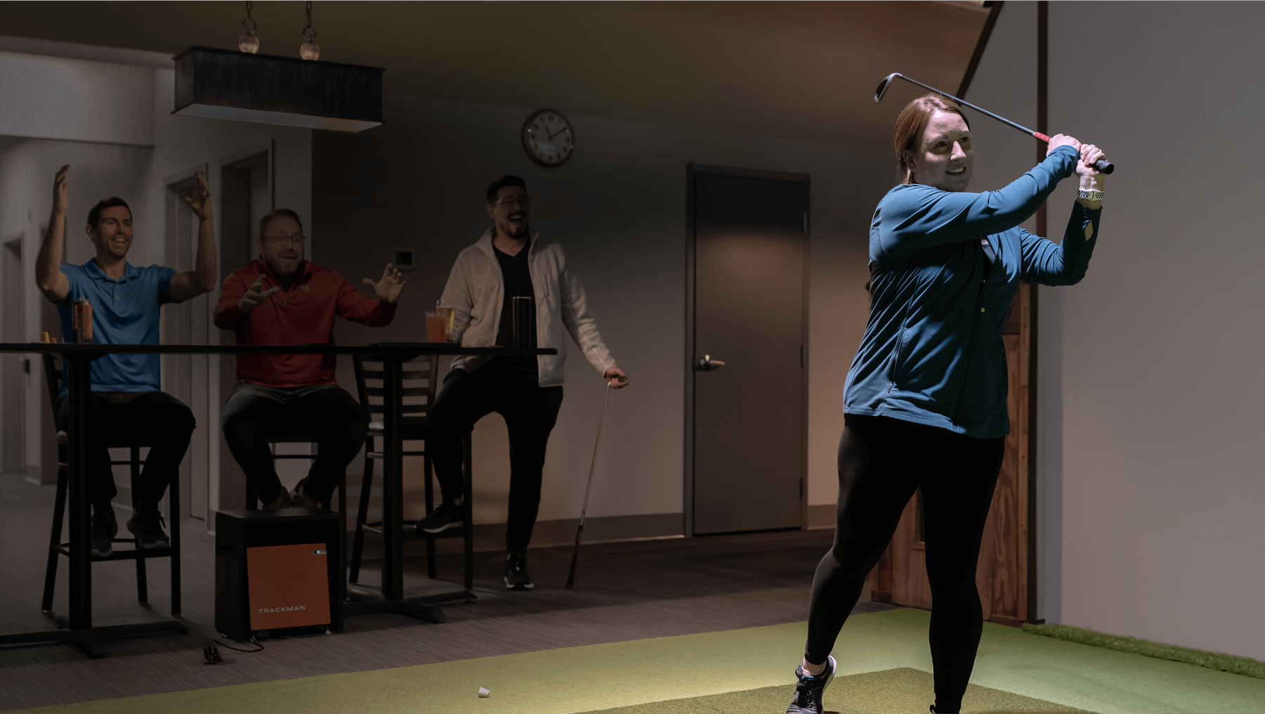 3 people watching a woman make a golf swing in a indoor golf setting. The people are happy and celebratory.