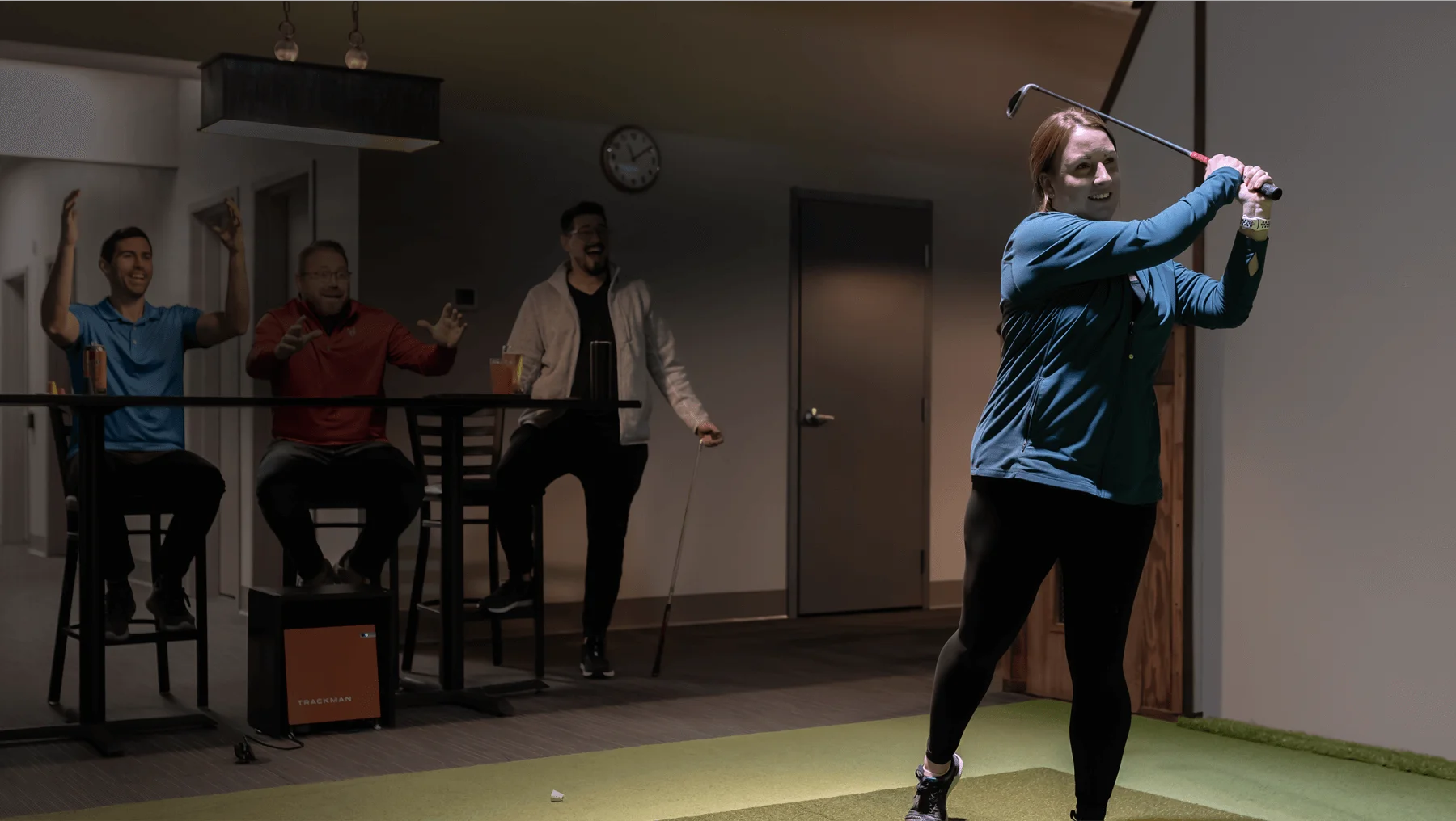3 people watching a woman make a golf swing in a indoor golf setting. The people are happy and celebratory.
